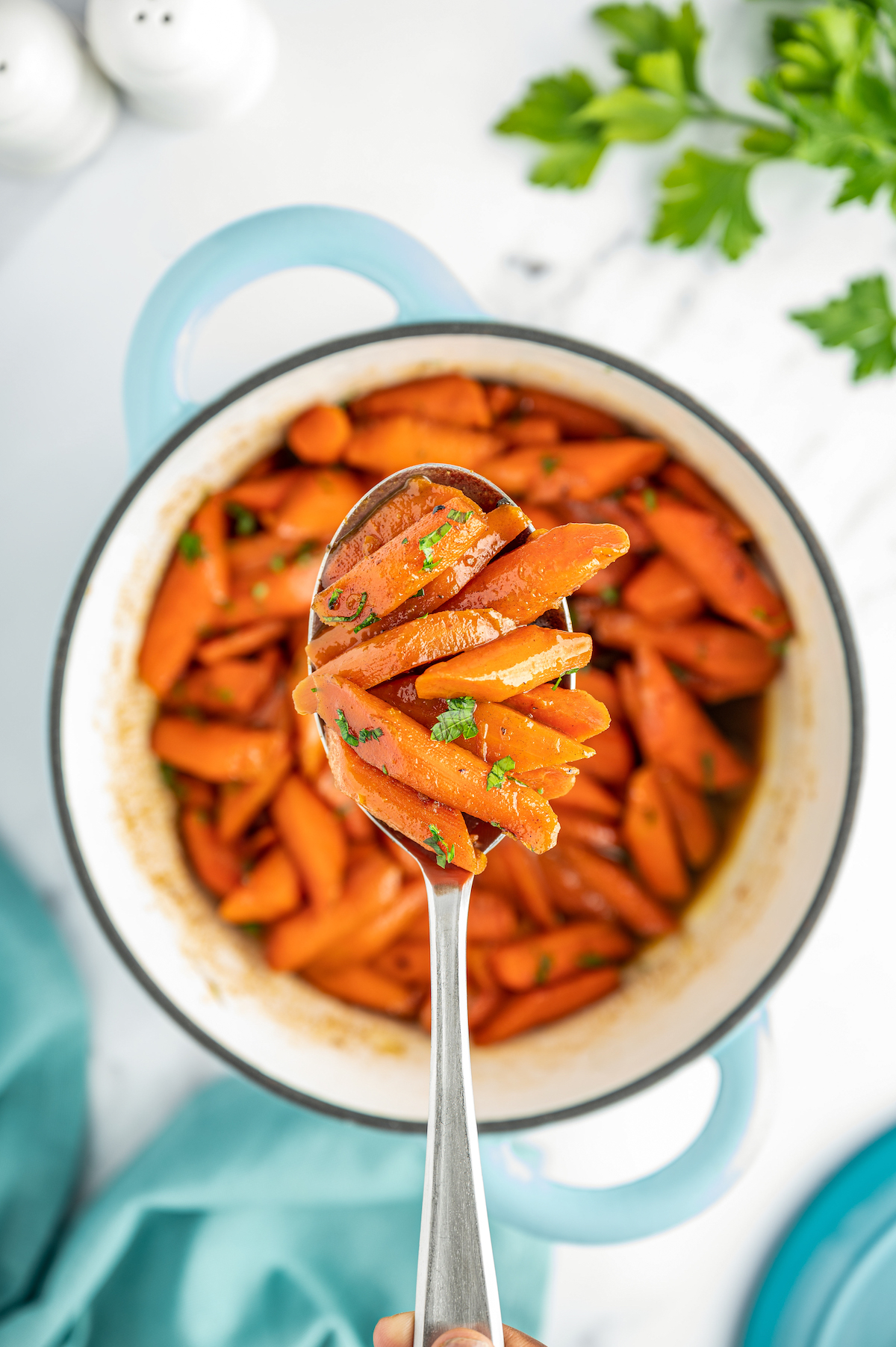 A pot of carrots in sauce, garnished with herbs. Some of the carrots are being lifted toward the camera on a spoon.