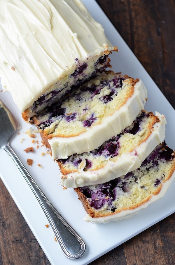 Overhead view of a Blueberry Pound Cake with cream cheese frosting on top sliced into pieces with a spatula next to it on a white platter.