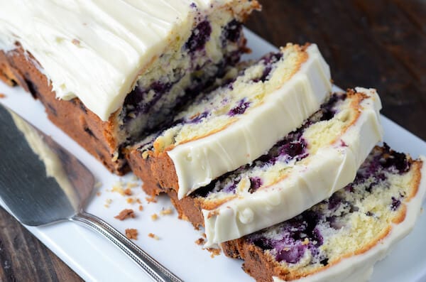 Blueberry Pound Cake with cream cheese frosting on top sliced into pieces with a spatula next to it on a white platter.