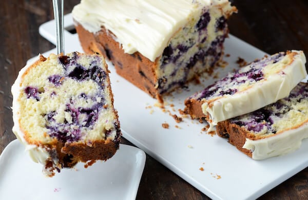 A slice of pound cake being served onto a white plate from a platter with a large blueberry lime pound cake sitting on top. 