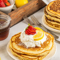A serving of lemon ricotta pancakes, garnished with fresh fruit and whipped cream, on a breakfast table with syrup and other items.