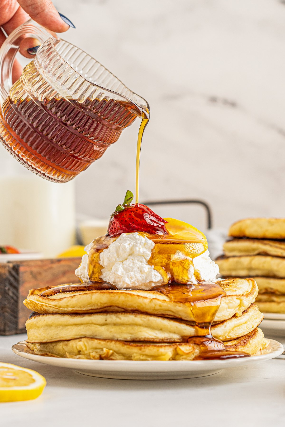 Three pancakes on a plate, topped with whipped cream and fruit. Syrup is being poured over the pancakes.