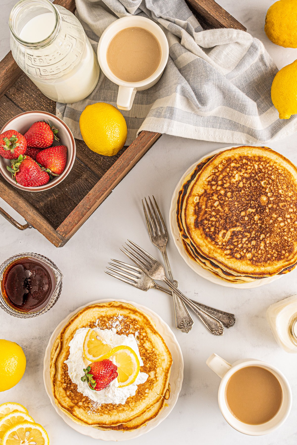 A table with milk, strawberries, lemons, coffee, cloth napkins, syrup, and a platter of homemade pancakes. Some of the pancakes have been placed and garnished on a smaller plate.