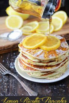 Maple syrup is poured over a plate of piled up pancakes with sliced lemons.
