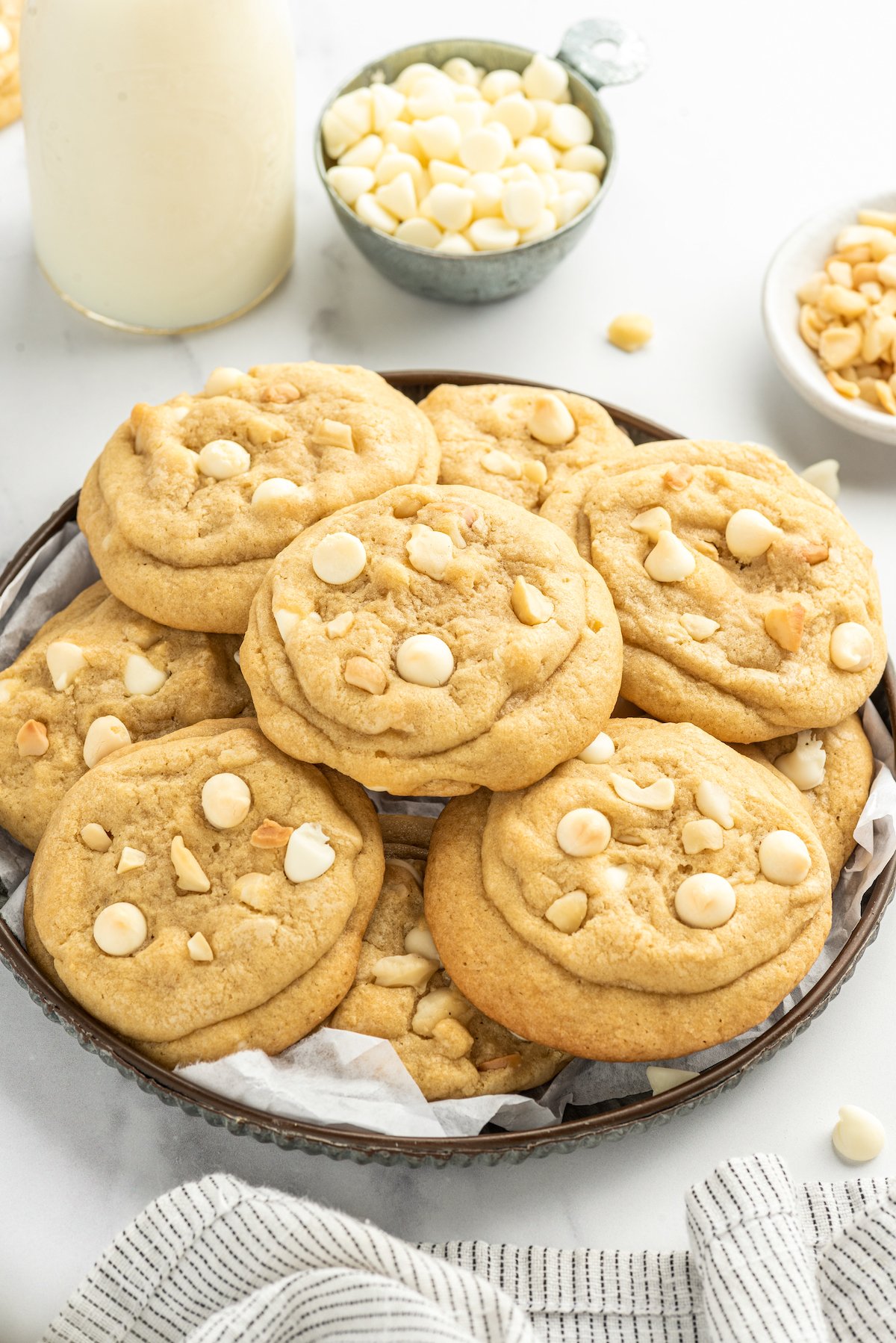 White chocolate cookies on a serving plate, with white chocolate chips and chopped nuts in small dishes nearby.