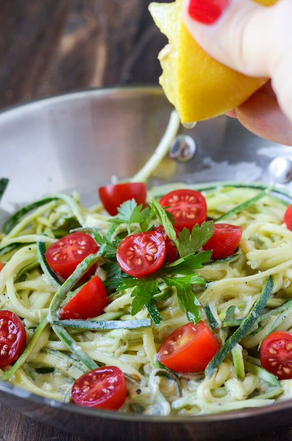 A lemon is squeezed over a bowl of creamy zoodles and cherry tomatoes.