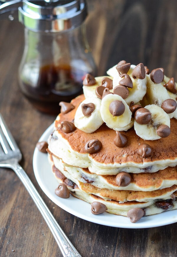 Three pancakes stacked on a plate with banana slices and chocolate chips piled on top in front of a maple syrup container.