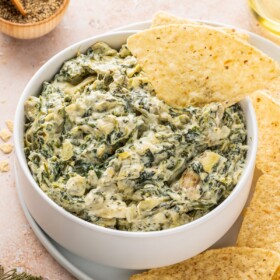 A white bowl of spinach artichoke dip with a tortilla chip in it.