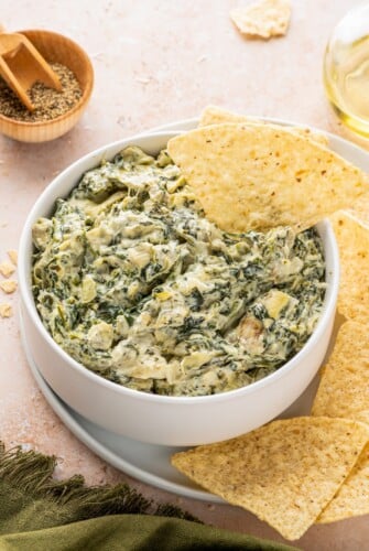 A white bowl of spinach artichoke dip with a tortilla chip in it.