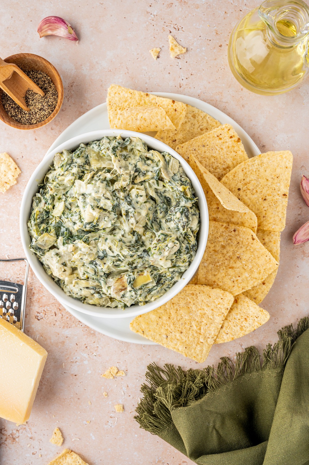 A bowl of dip with a plate of chips on a table with a cloth napkin, salt, pepper, and other items.