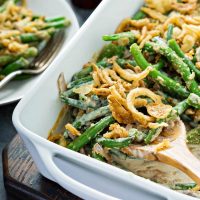 Green Bean Casserole topped with french fried onions in a white baking pan with a wooden spoon removing a serving