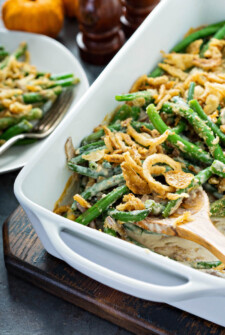 Green Bean Casserole topped with french fried onions in a white baking pan with a wooden spoon removing a serving