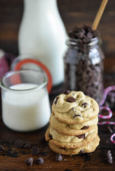A stack of classic chocolate chip cookies next to a glass of milk and jar of chocolate chips.
