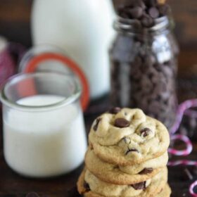 A stack of classic chocolate chip cookies next to a glass of milk and jar of chocolate chips.