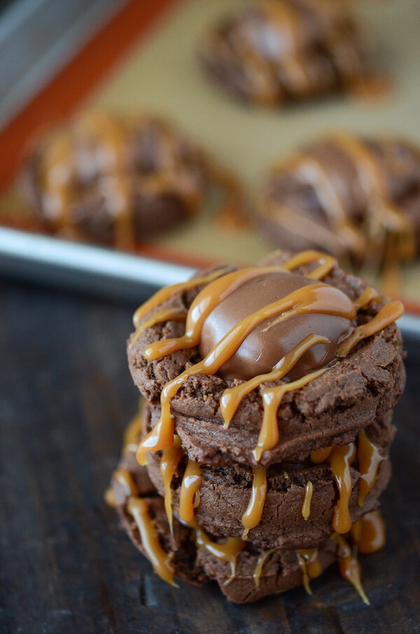 Stack of Turtle Chocolate Cookies topped with a caramel drizzle, with cookies on a baking sheet in the background.