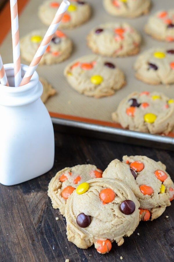 Three cookies with Reese's Pieces in them are piled in front of a baking sheet with more cookies