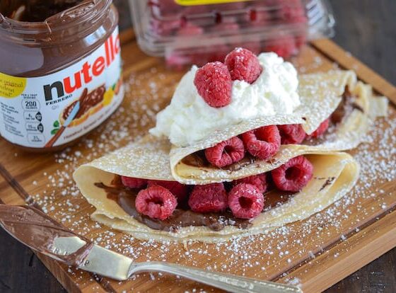 Raspberry Nutella Crepes topped with whipped cream and raspberries. Jar of Nutella in the background.