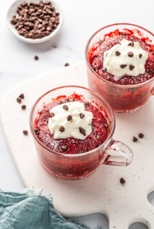 Two red velvet mug cakes topped with frosting and chocolate chips.