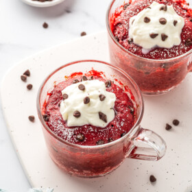 Two red velvet mug cakes topped with frosting and chocolate chips.