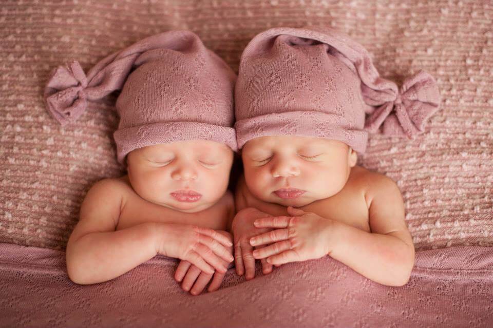 Newborn Twins Sleeping Tucked Into a Pink Blanket Wearing Blush Caps