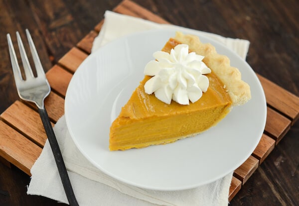 Buttermilk Pumpkin Pie topped with whipped cream on a white plate with a fork on a wooden board