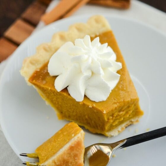 Buttermilk Pumpkin Pie topped with whipped cream on a white plate with a fork on a wooden board