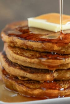 Stack of Pumpkin Pancakes topped with butter and being drizzled with syrup on a white plate