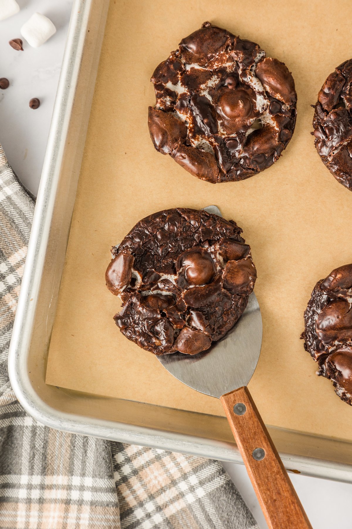 Lifting a cookie off of a baking sheet filled with chocolate chip marshmallow cookies with a spatula.