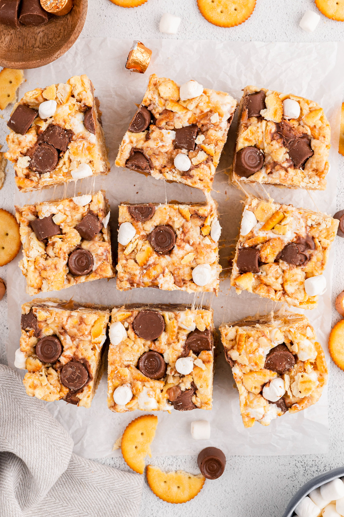 Overhead shot of nine marshmallow treat bars on a sheet of parchment, with Ritz crackers, Rolos, and marshmallow scattered around.