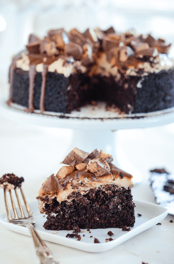 Slice of Reese's Dark Chocolate Peanut Butter Cake on a white plate with a fork topped with chopped Reese's Peanut Butter Cups over chocolate ganache - remainder of cake in the background