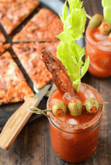 Bacon Bloody Mary with bacon, a celery stalk, and green olive skewers - pizza is shown in the background