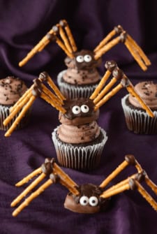 Two Pretzel Spiders on Top of Cupcakes and One on its Own on Top of a Purple Tablecloth