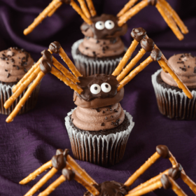 Two Pretzel Spiders on Top of Cupcakes and One on its Own on Top of a Purple Tablecloth