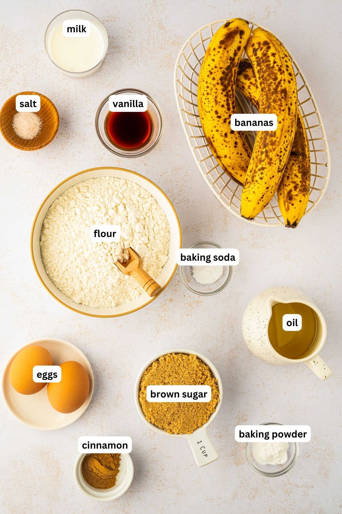 Ingredients arranged in bowls for this easy banana muffin recipe. From top to bottom: milk, salt, vanilla, over-ripe bananas, flour, baking soda, oil, eggs, brown sugar, baking powder and cinnamon.