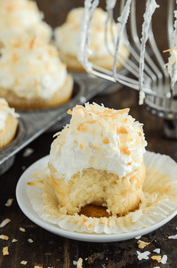 Triple Coconut Poke Cupcakes piled high with whipped cream and shredded coconut, with a bite missing.