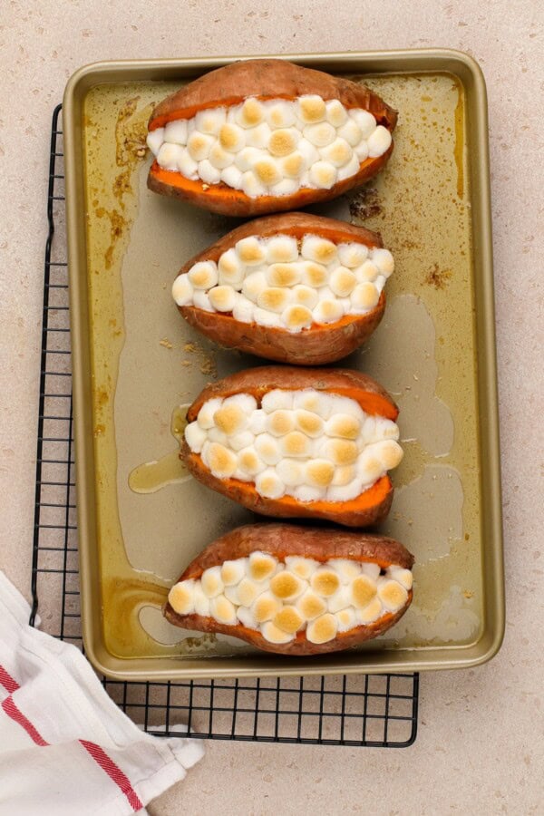 Four sweet potatoes lined up on a baking dish with toasted marshmallows.