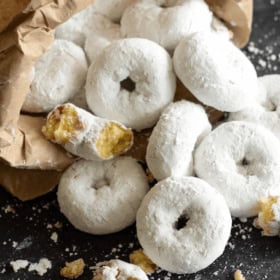 A Pile of Homemade Doughnuts Covered in Confectioner's Sugar with a Paper Bag Holding Additional Doughnuts