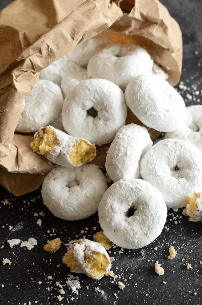 A Pile of Homemade Doughnuts Covered in Confectioner's Sugar with a Paper Bag Holding Additional Doughnuts