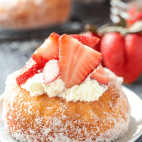 A Strawberry Shortcake Donut on a White Plate with Whipped Cream and Strawberry Slices on Top