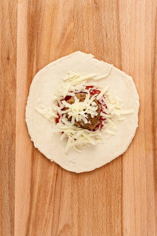 A flattened biscuit round is topped with marinara, shredded cheese, and a single meatball.