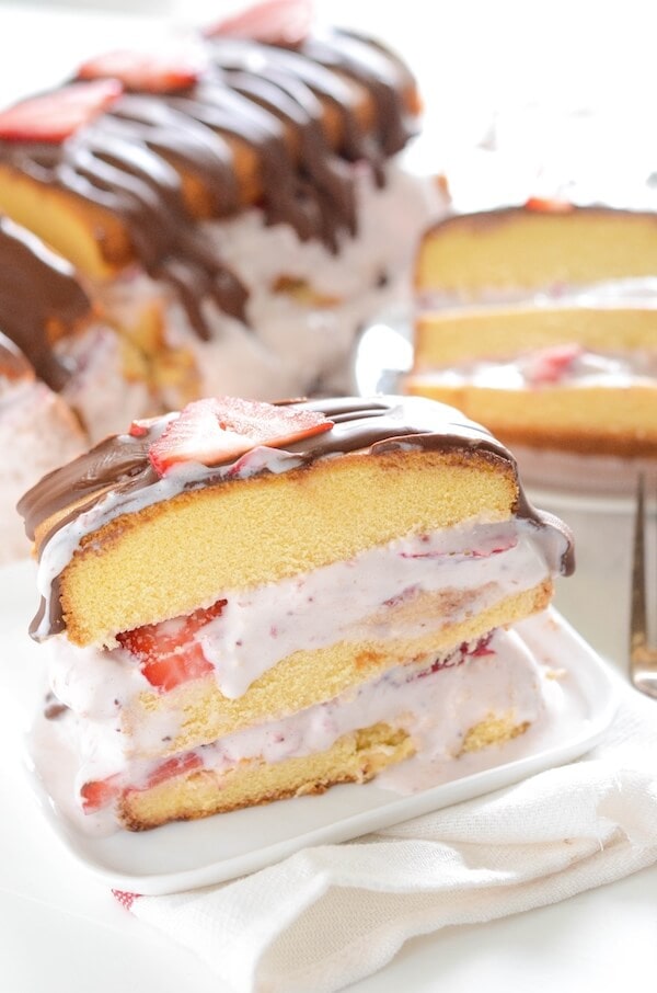 A slice of strawberry ice cream cake on a plate with the rest of the cake in the background.