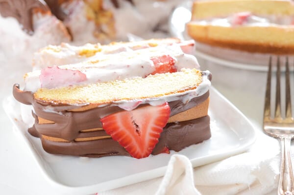 A slice of strawberry ice cream cake on a plate with the rest of the cake in the background.