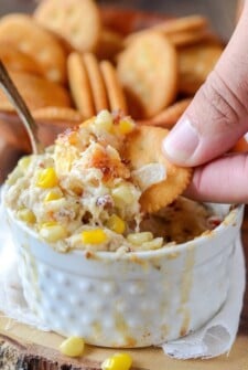 Hot Crab, Corn and Bacon Dip in a white baking dish with someone dipping a cracker