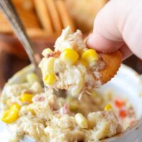 Hot Crab, Corn and Bacon Dip in a white baking dish with someone dipping a cracker