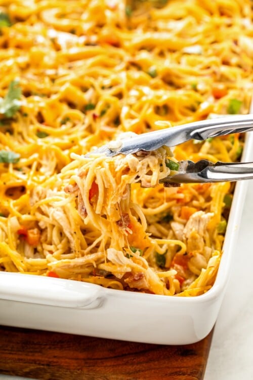 Metal tongs are lifting a portion of creamy, cheesy pasta from a baking dish.