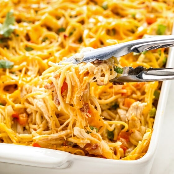 Metal tongs are lifting a portion of creamy, cheesy pasta from a baking dish.