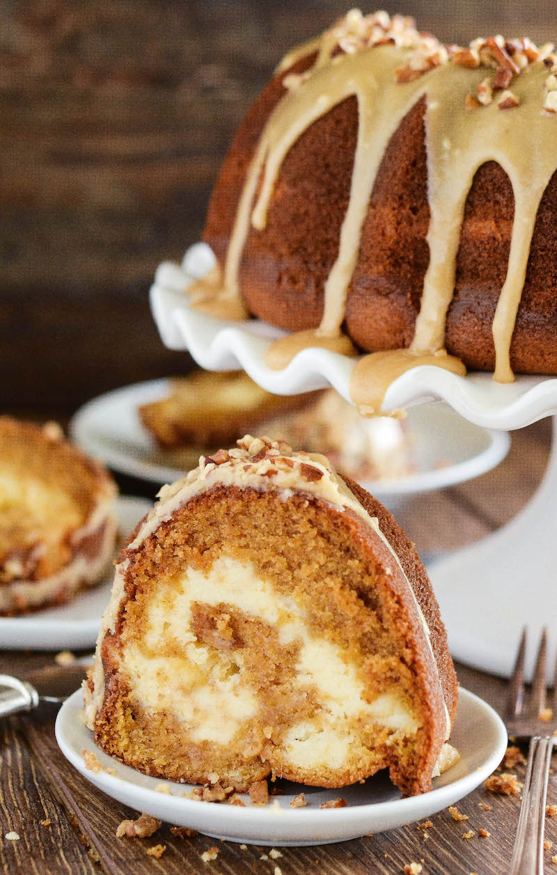 A slice of sweet potato cake with a cream cheese swirl, on a white plate with the rest of the cake on a cake stand in the background.