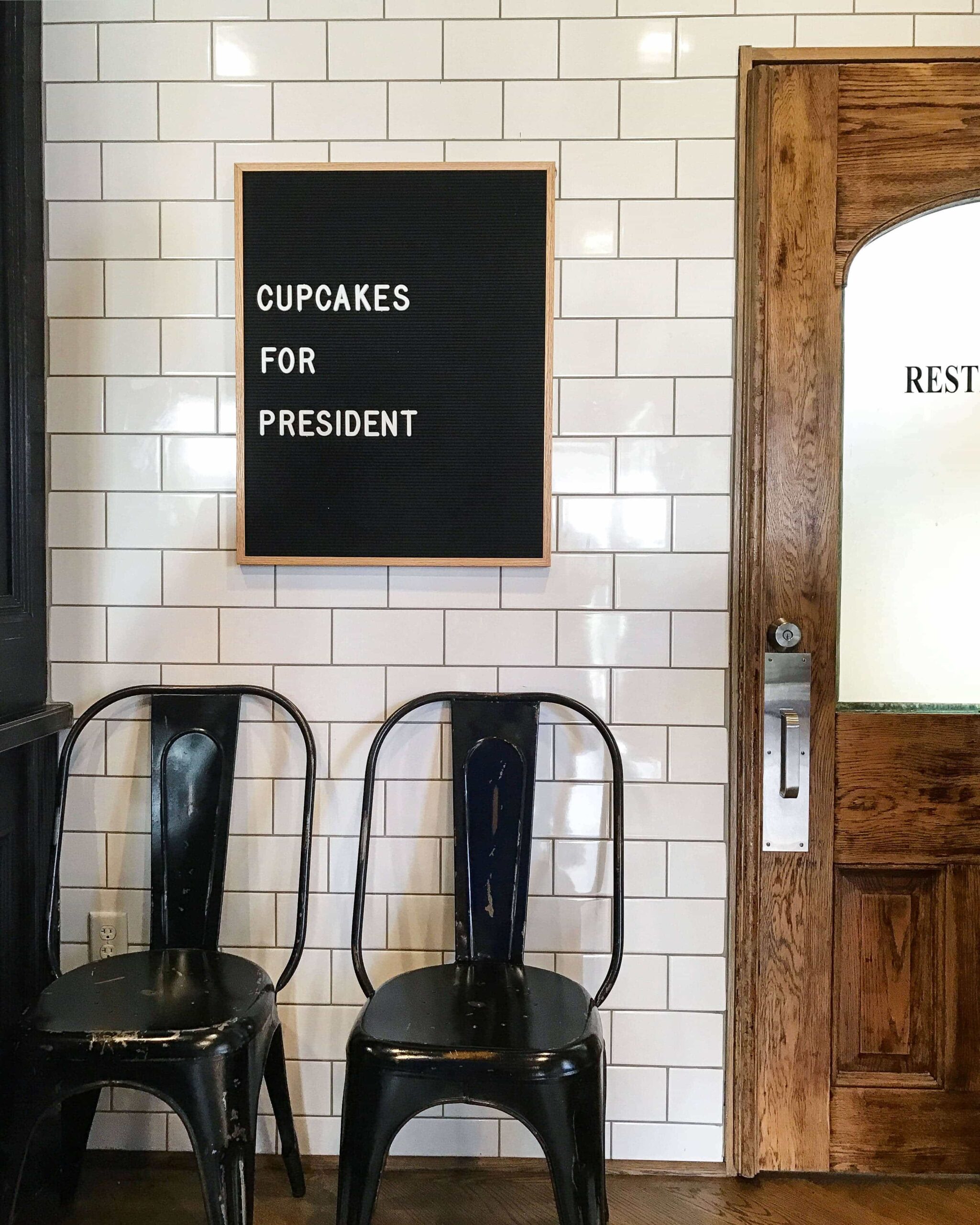 A Sign on a Shiplap Wall Beside a Restroom that Says "Cupcakes for President"
