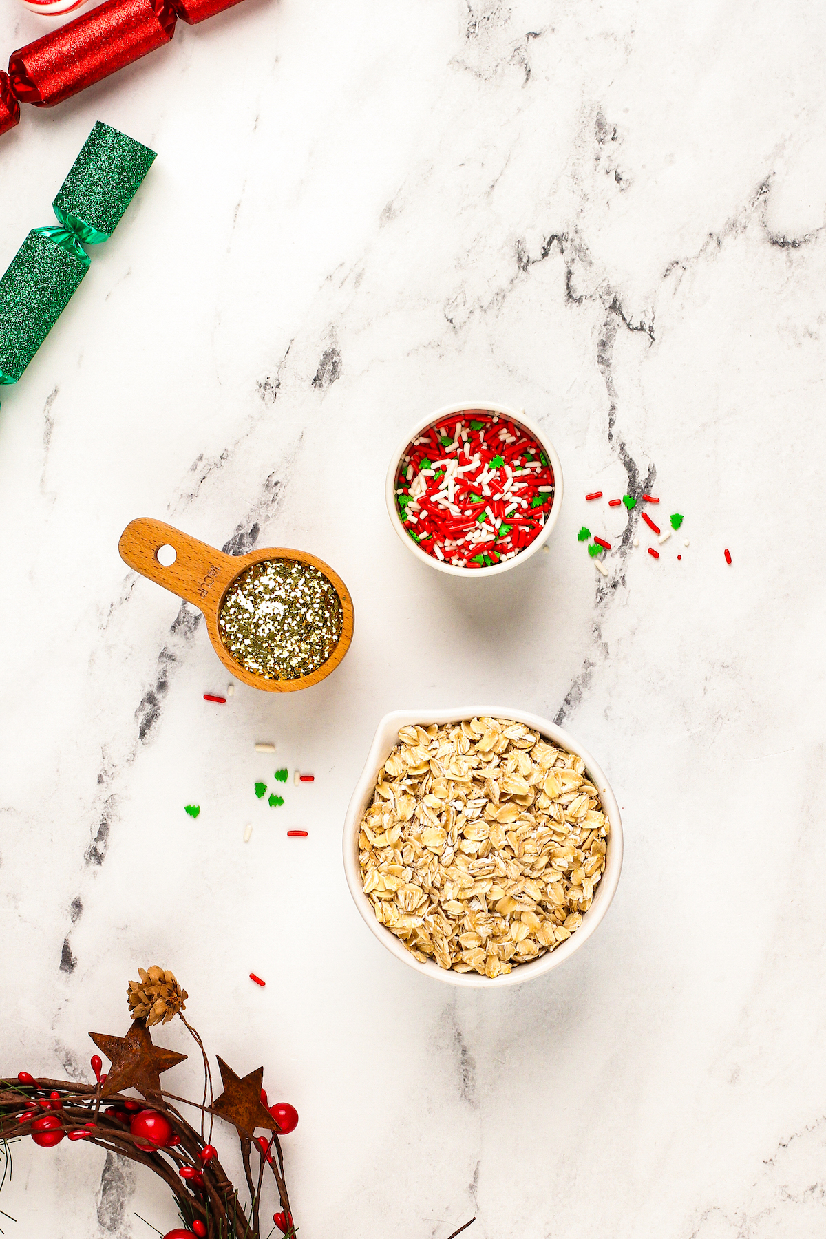 Ingredients and materials for reindeer food recipe arranged in bowls. From top to bottom: a bowl of festive colored sprinkles, a measuring cup of gold glitter and a bowl of oats.