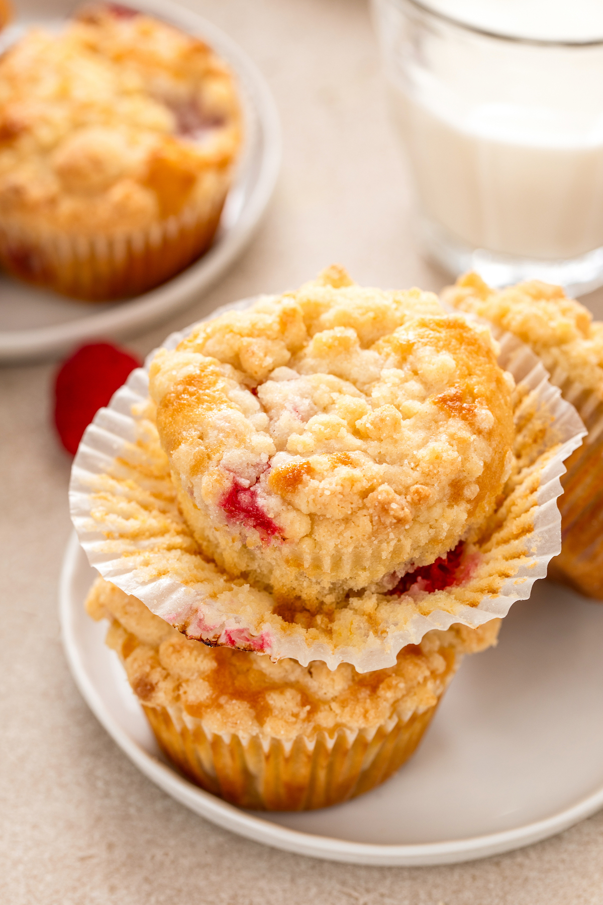 Moist and tender raspberry muffins with sweet crumb topping stacked on top of each other with the paper liner on the top muffin being pulled off.